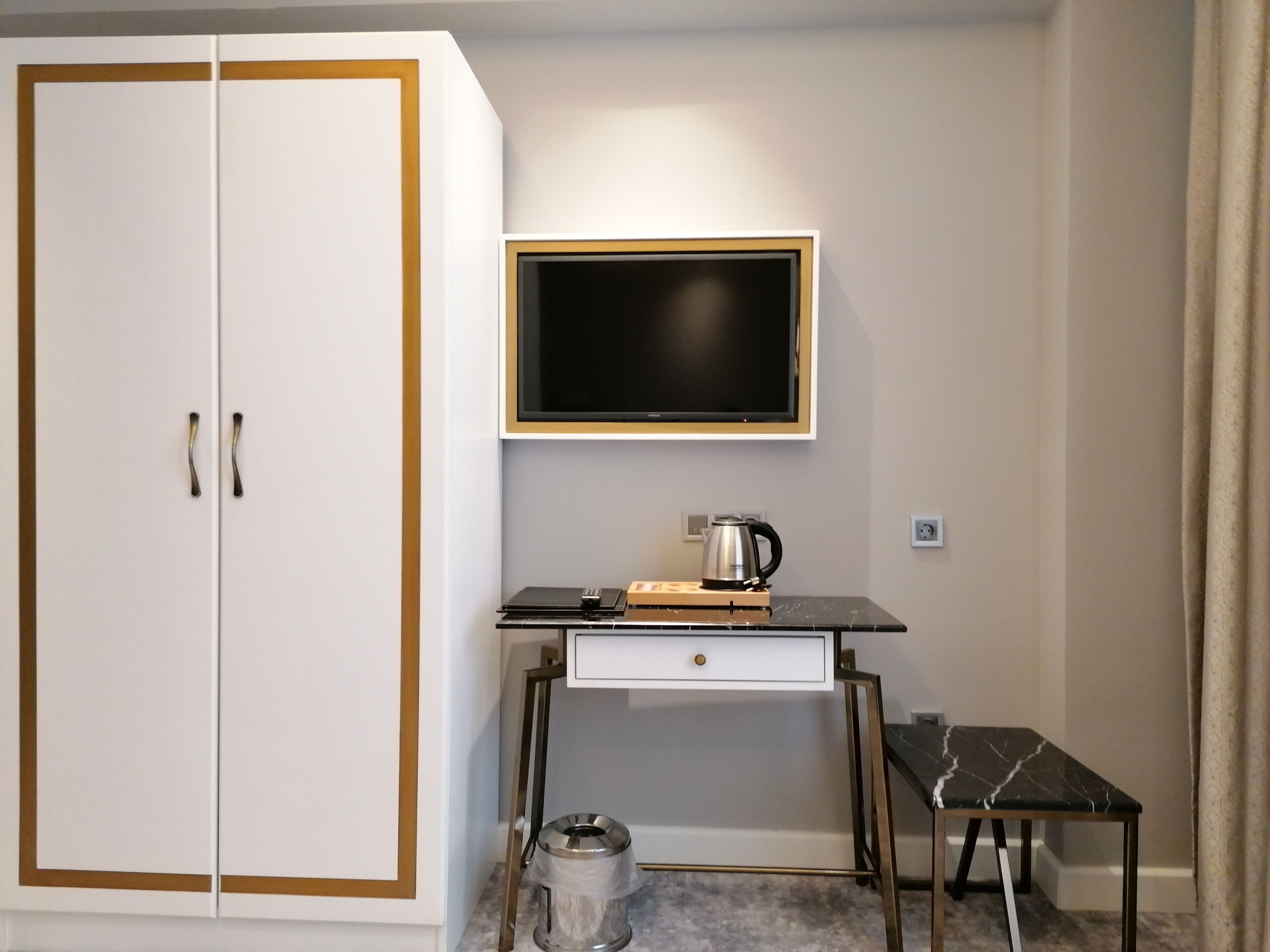 A room with a white wardrobe, a desk with a television, a kettle, and a trash bin.