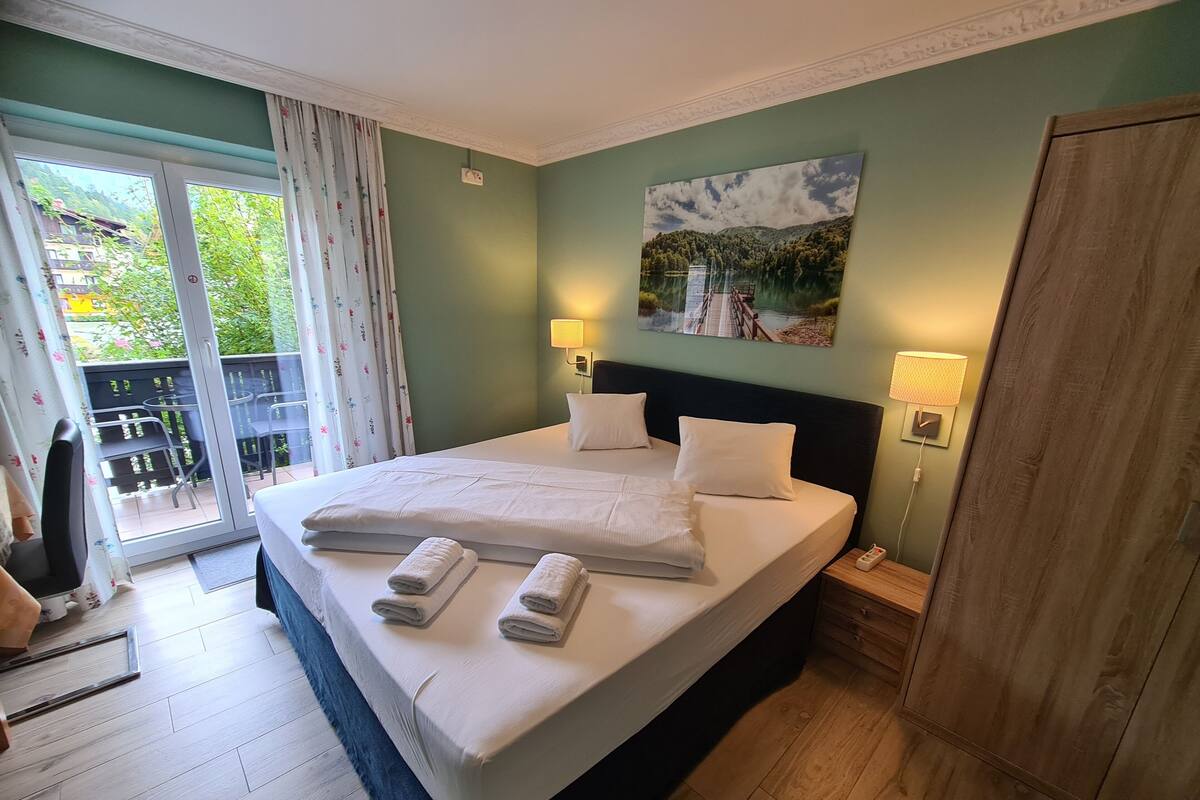 A hotel room with a large bed, two bedside lamps, a painting on the wall, and a view of a balcony with outdoor furniture.