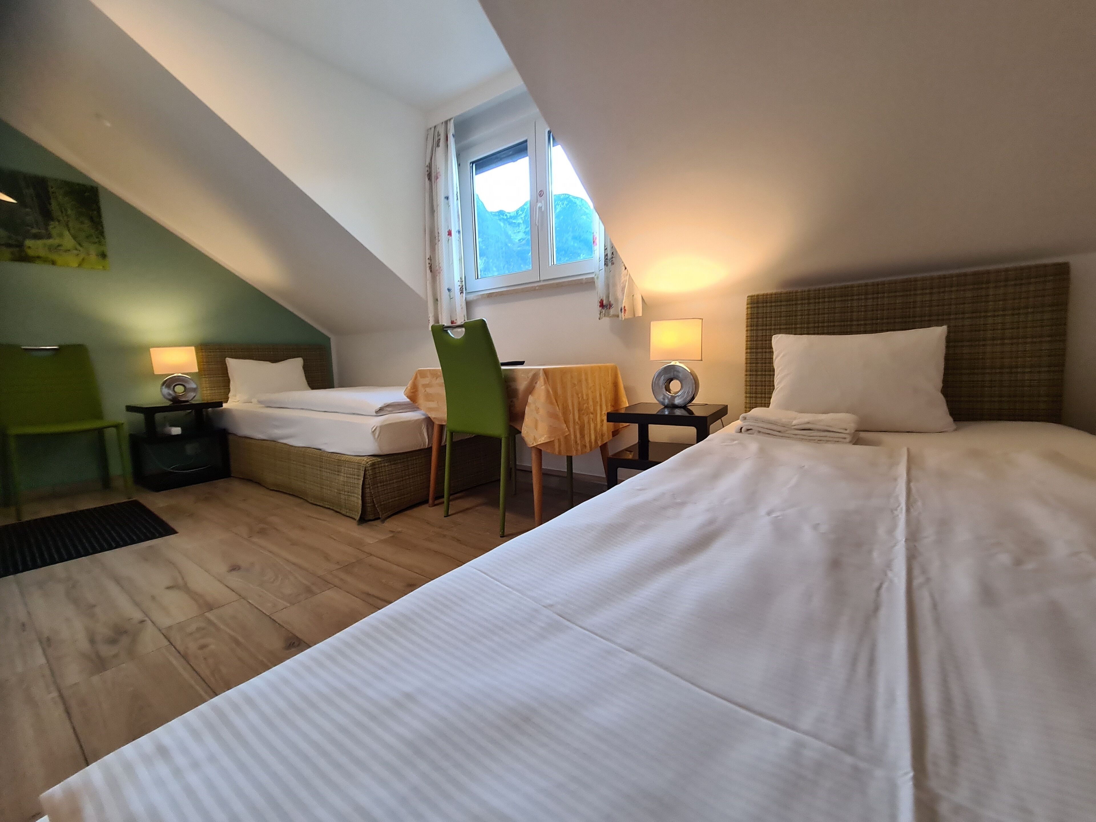 Habitación de hotel con dos camas, una mesa de comedor, una silla, una mesita con una lámpara y una ventana con vistas a la montaña.
