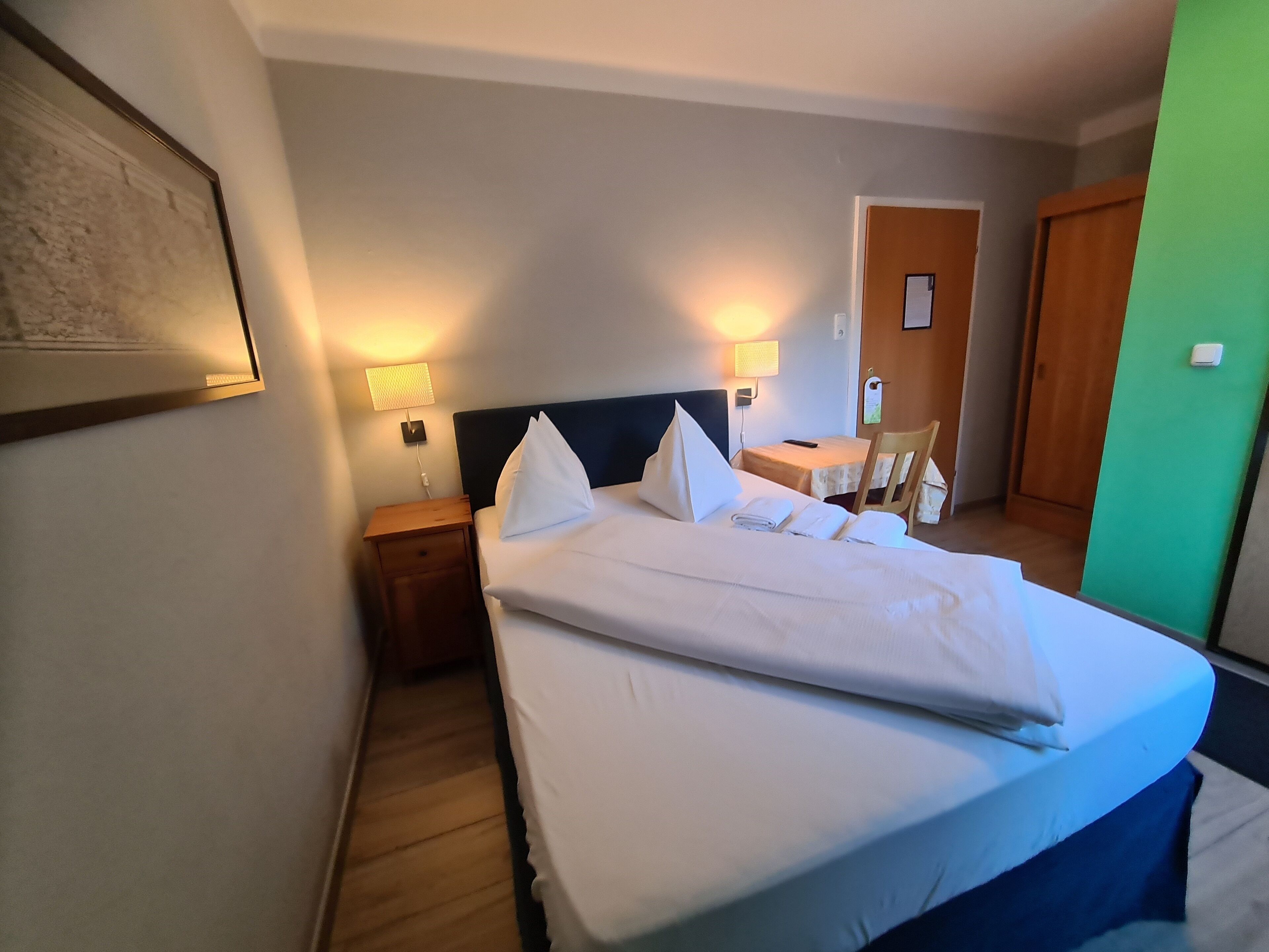 A hotel room with a neatly made bed, bedside tables with lamps, a small table with a chair, and a door.