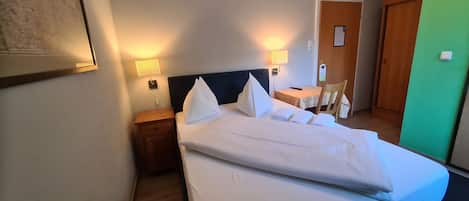 A hotel room with a neatly made bed, bedside tables with lamps, a small table with a chair, and a door.