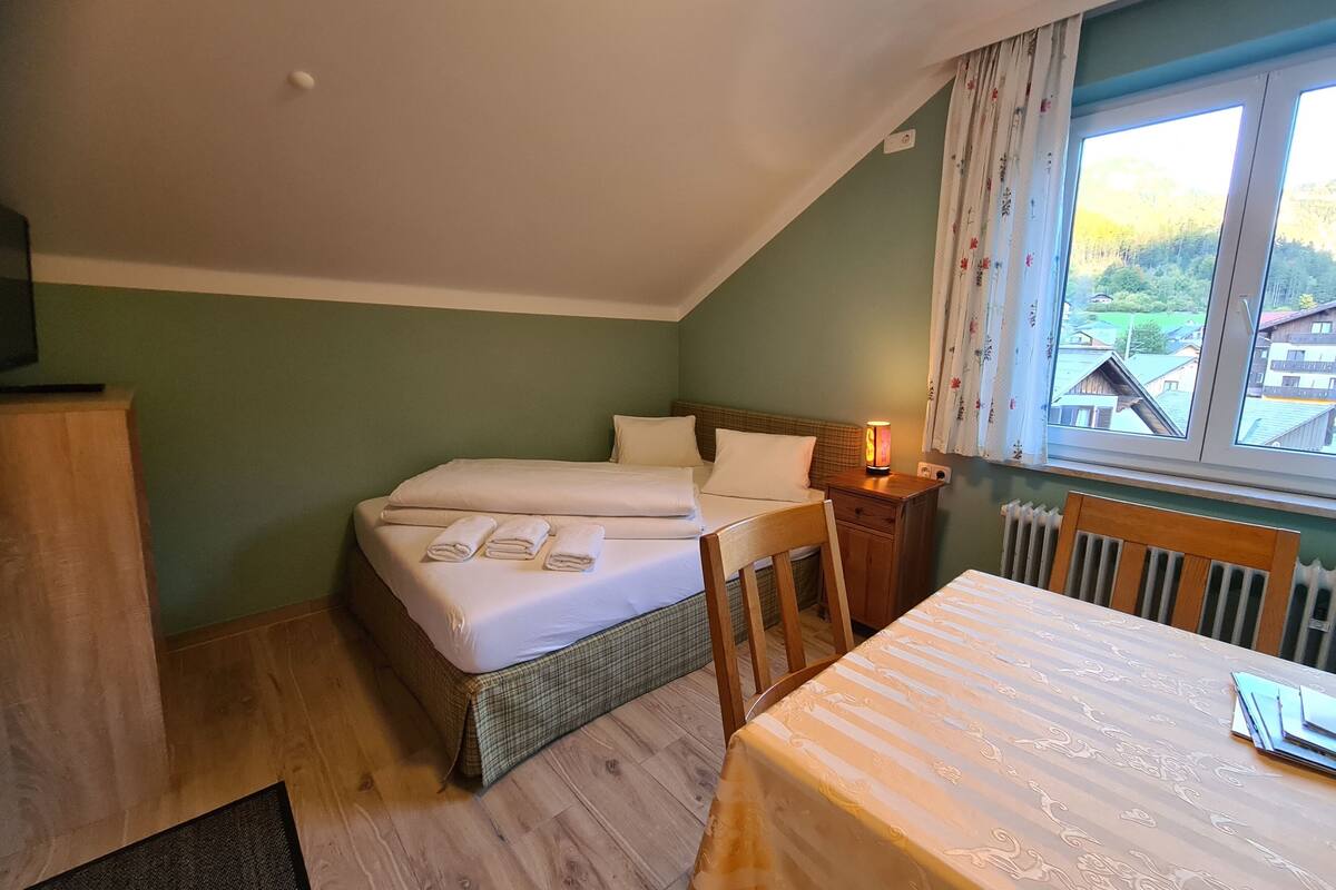A bedroom with a bed, a table, a chair, a nightstand, and a window with a view of houses.