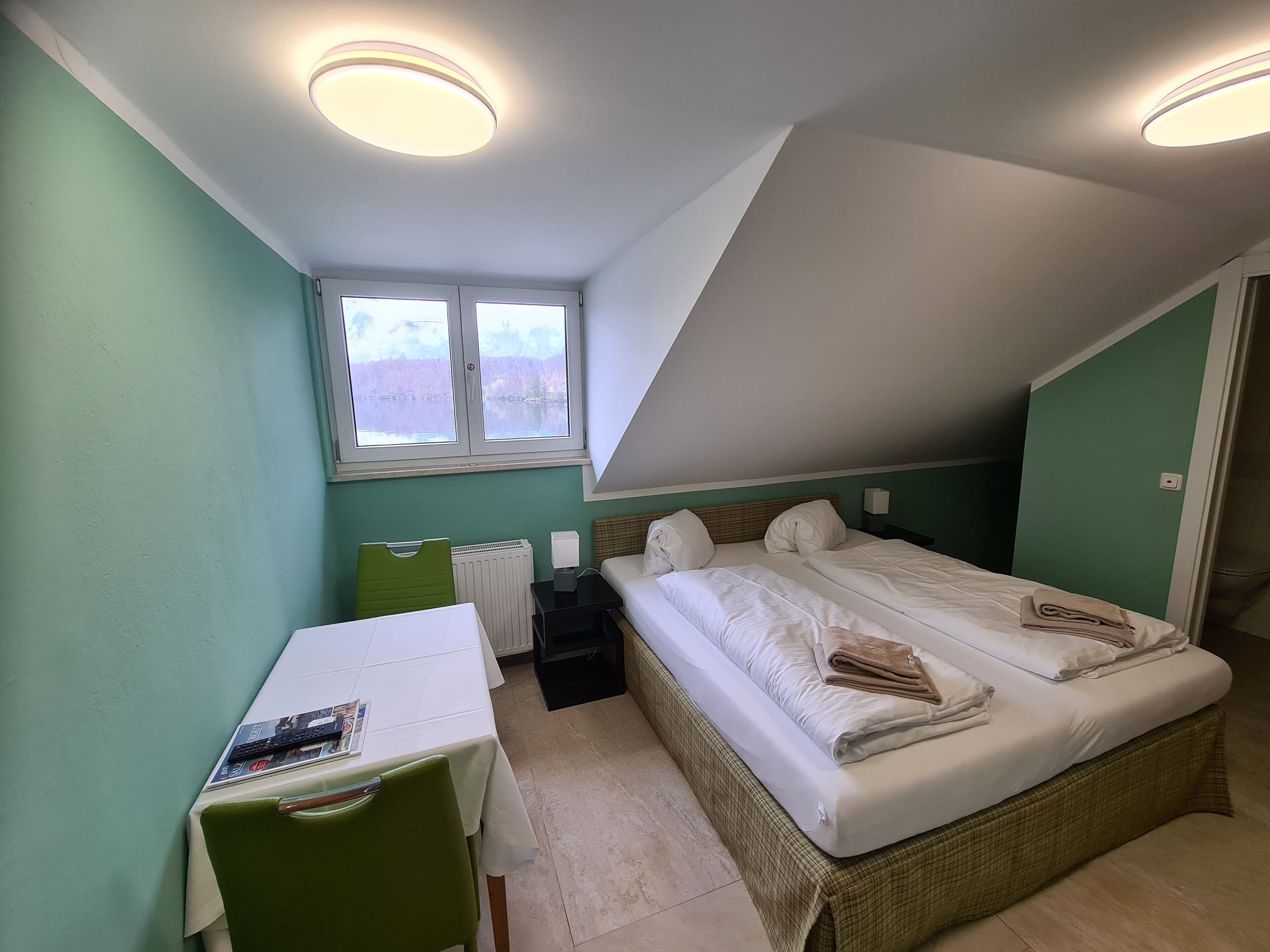 A small, well-lit room with a bed, a desk, and a chair. There is a window providing natural light and a view of the outdoors.