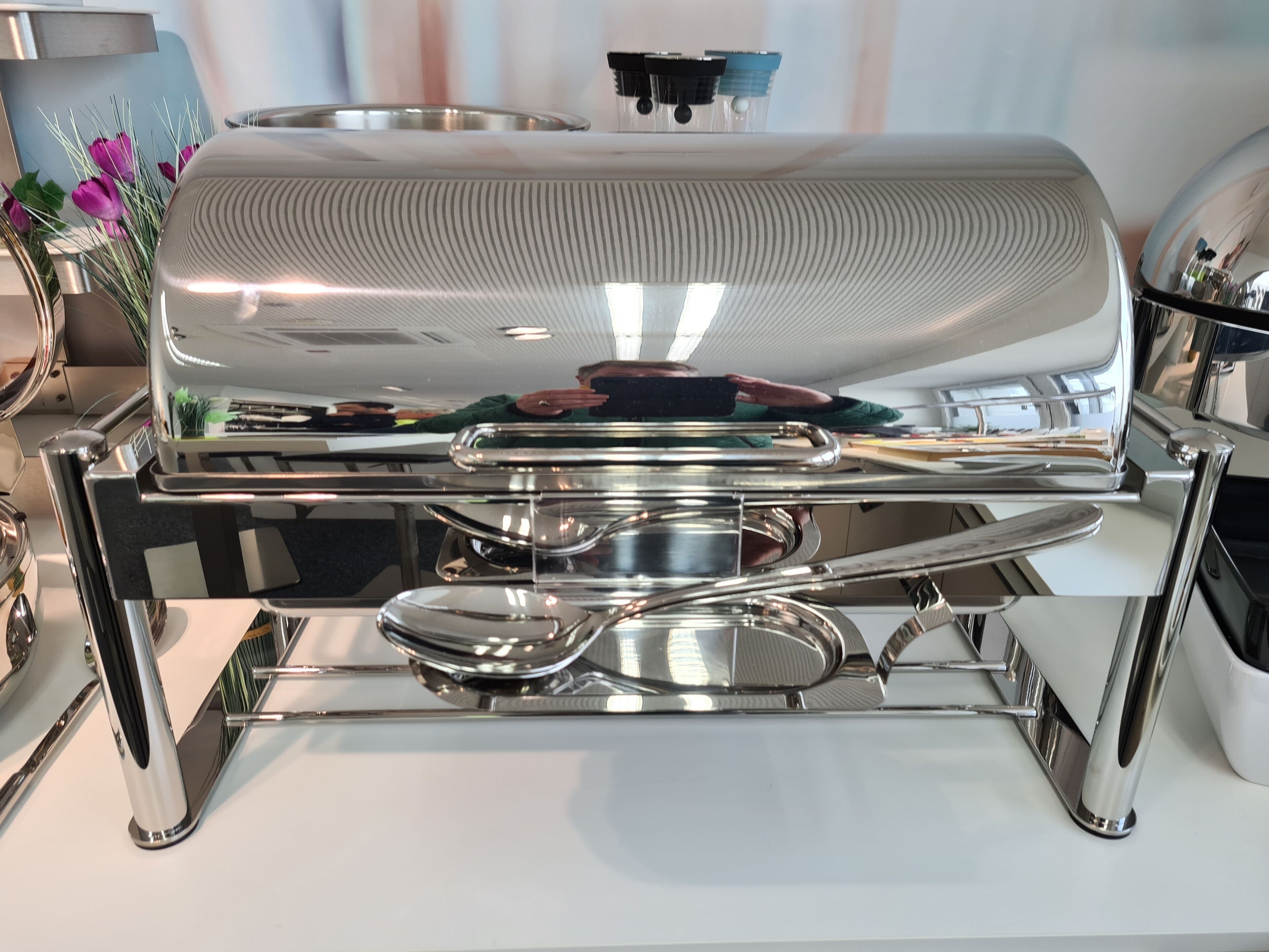 Una mesa de buffet de acero inoxidable con una parrilla grande, bandejas de servicio y un dispensador de agua.