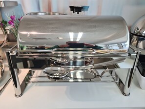 Una mesa de buffet de acero inoxidable con una parrilla grande, bandejas de servicio y un dispensador de agua.