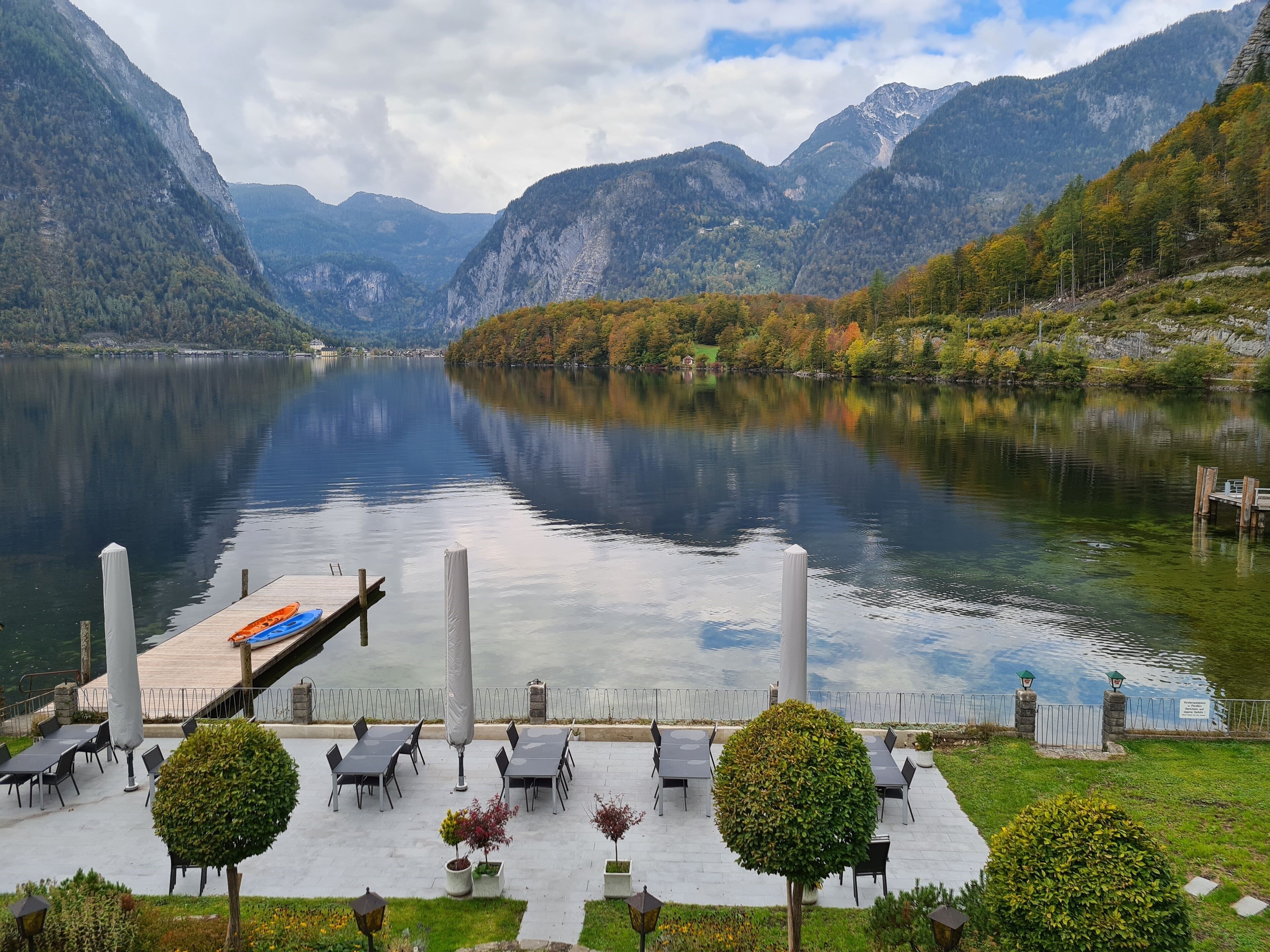 Un patio junto al lago con mesas y sillas, rodeado de montañas y un muelle con kayaks.
