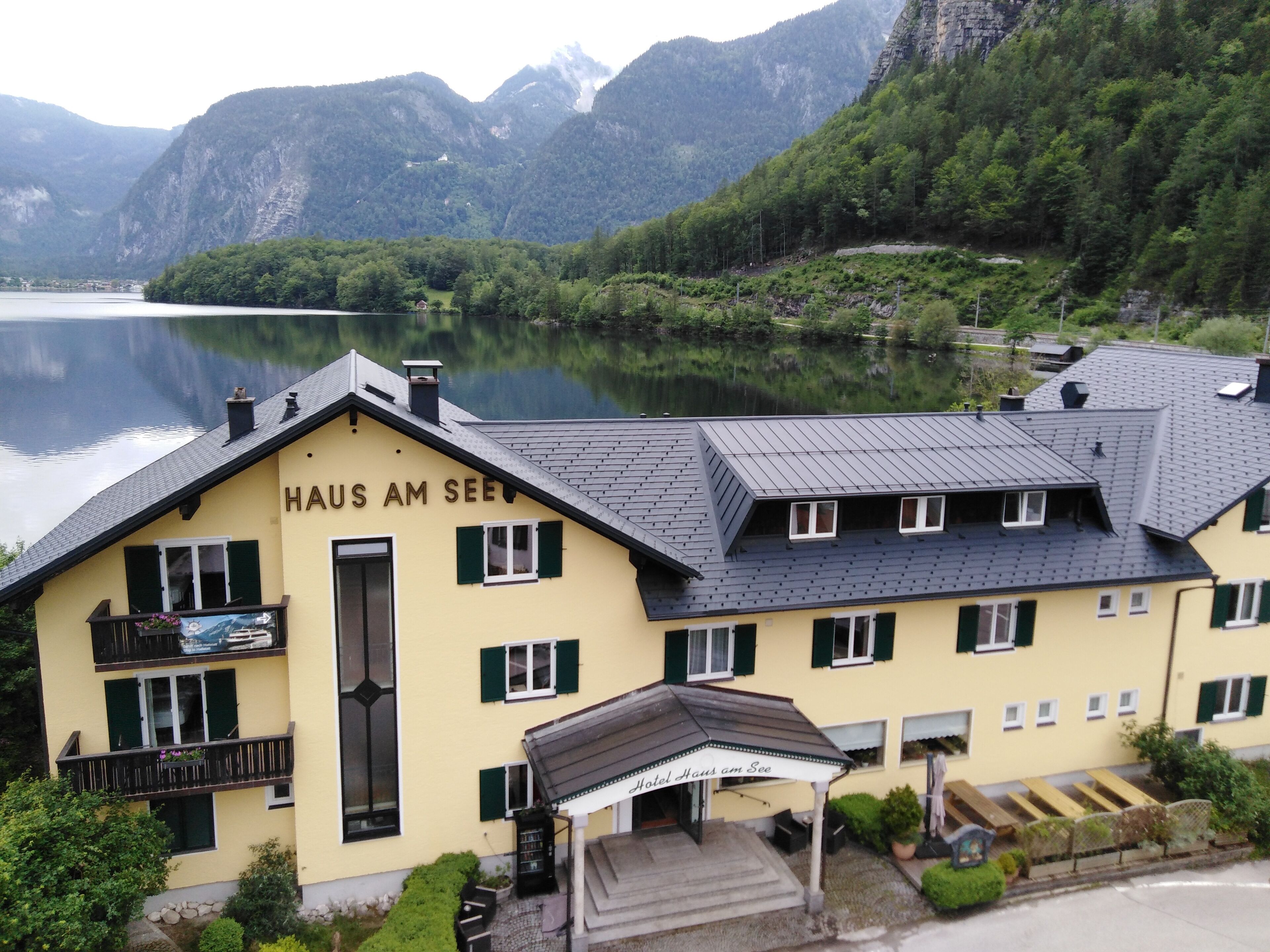 A yellow building with the sign "HAUS AM SEE" indicating a hotel. It features a balcony with flowers, a covered entrance, and a view of a lake and mountains in the background.