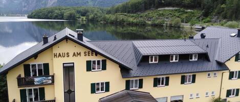 A yellow building with the sign "HAUS AM SEE" indicating a hotel. It features a balcony with flowers, a covered entrance, and a view of a lake and mountains in the background.
