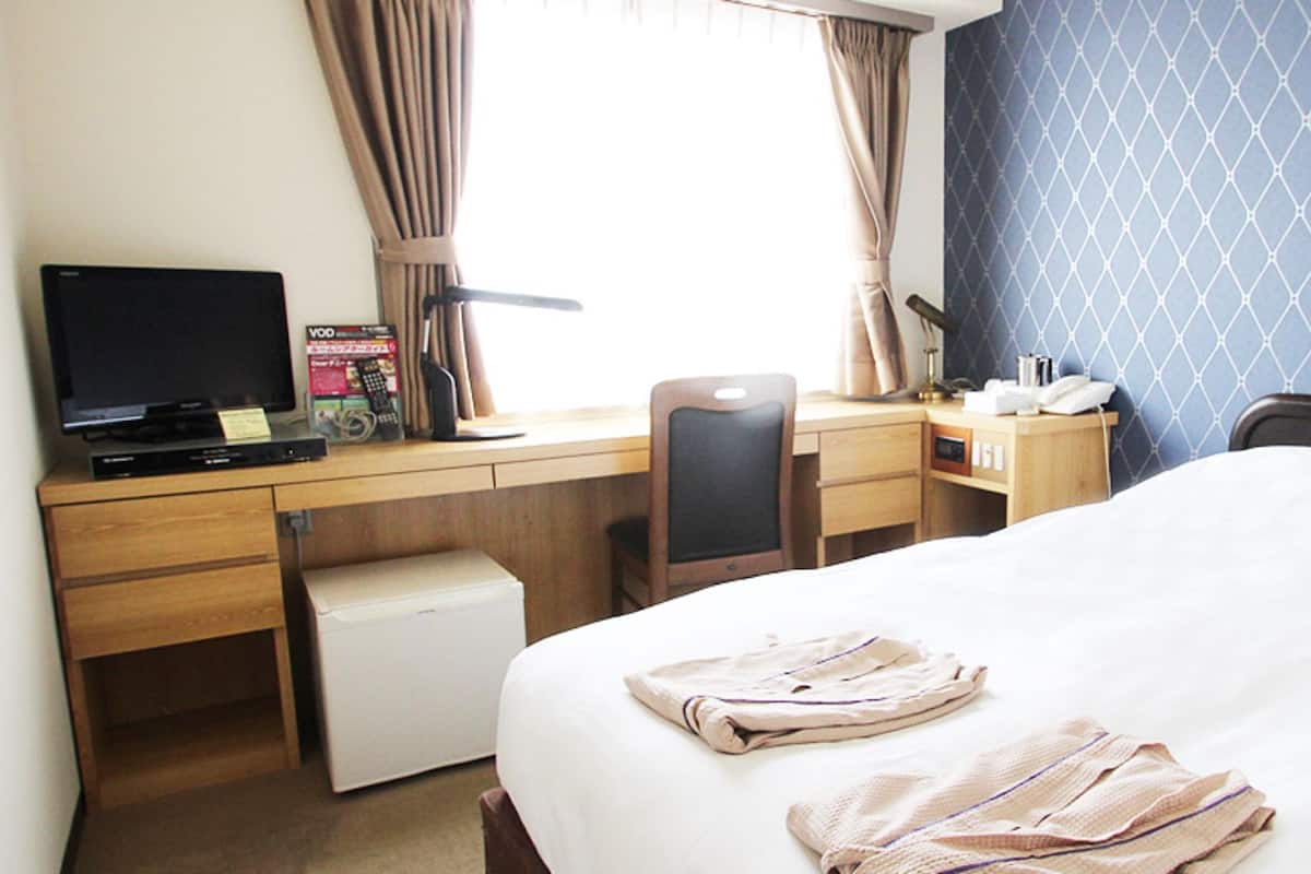 A hotel room with a bed, a desk with a computer, a chair, a small fridge, and a window with curtains.