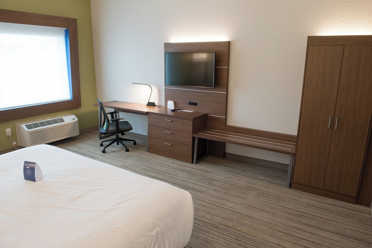 A hotel room with a bed, desk, chair, TV, wardrobe, and air conditioning unit.