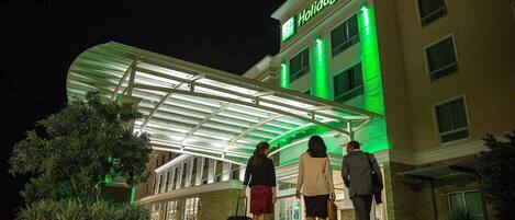 Entrada de un hotel con un vestíbulo techado, fachada de vidrio y un cartel que dice 'Holiday Inn'.