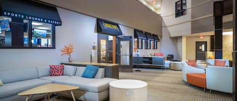 A modern hotel lobby with a sports lounge and eatery, a reception desk, seating areas, and a patterned carpet.