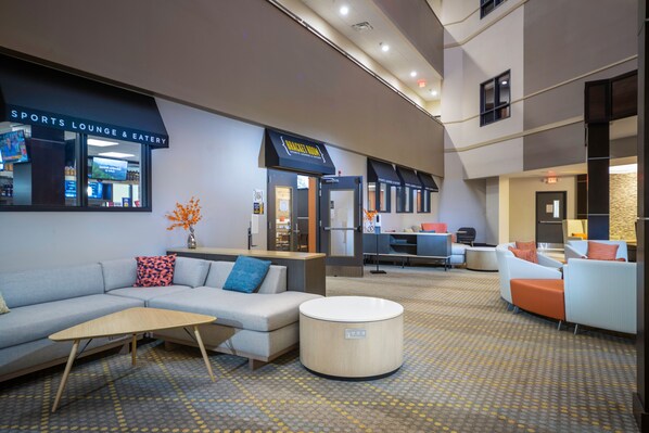 Un hall d’hôtel moderne comprenant un bar sportif avec espace de restauration, un comptoir d’accueil, des coins salon et un tapis à motifs.