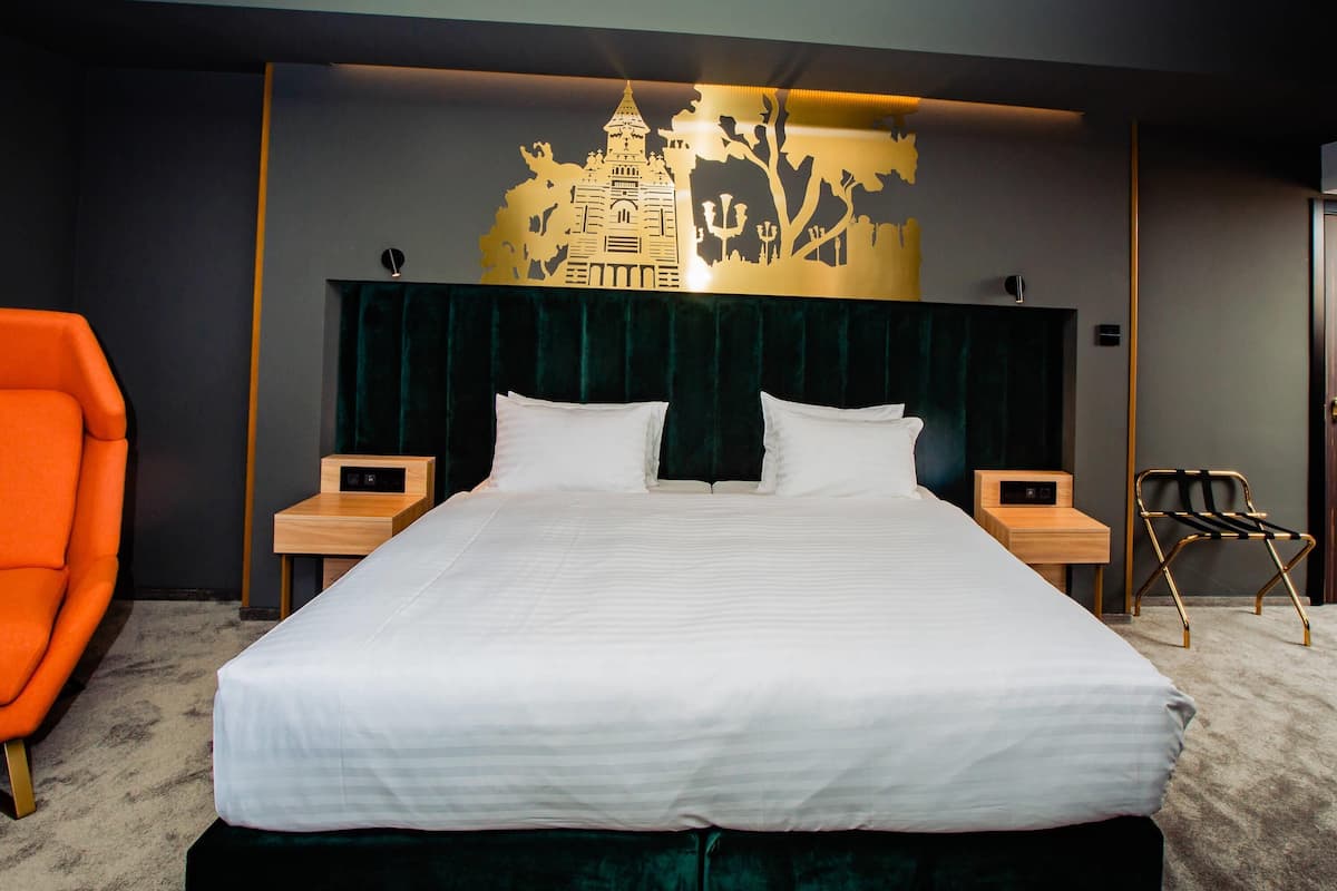 A modern hotel room with a large bed, two bedside tables, a nightstand, a chair, and a decorative wall piece.
