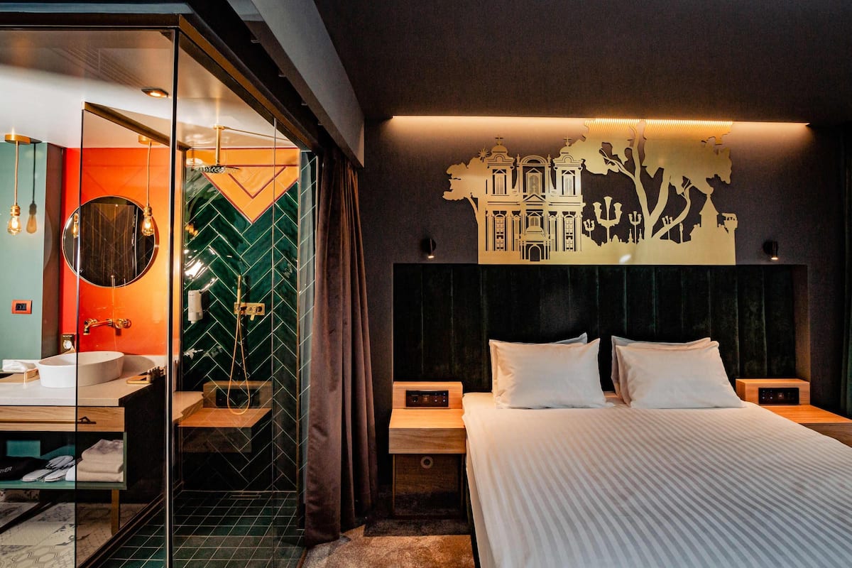 A modern hotel room with a large bed, a separate bathroom with a glass shower, and a cityscape mural on the wall.