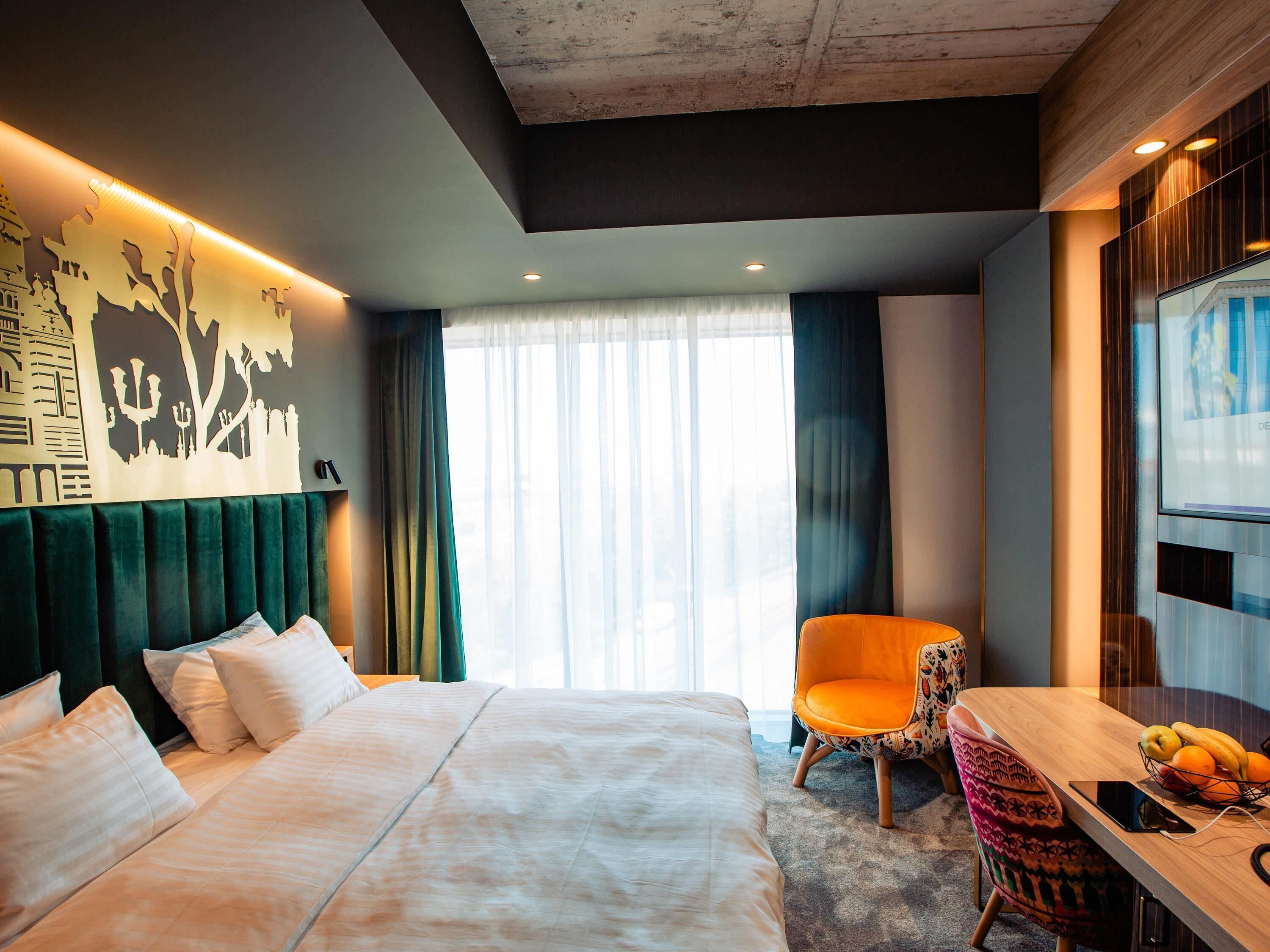 A hotel room with a bed, a desk with a chair, a television, and a large window with curtains.