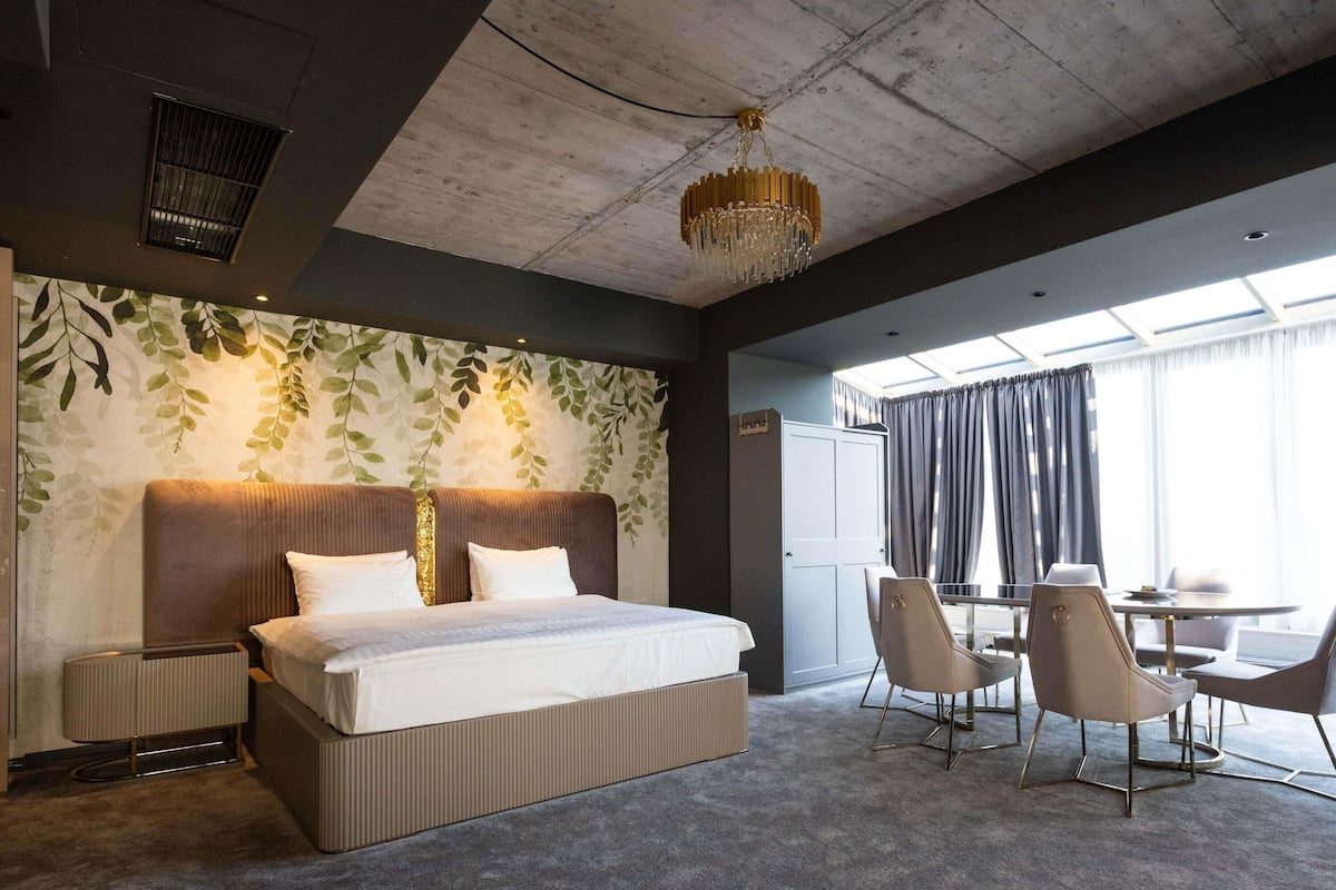 A modern hotel room with a large bed, a desk with chairs, a chandelier, and a view of the outdoors.