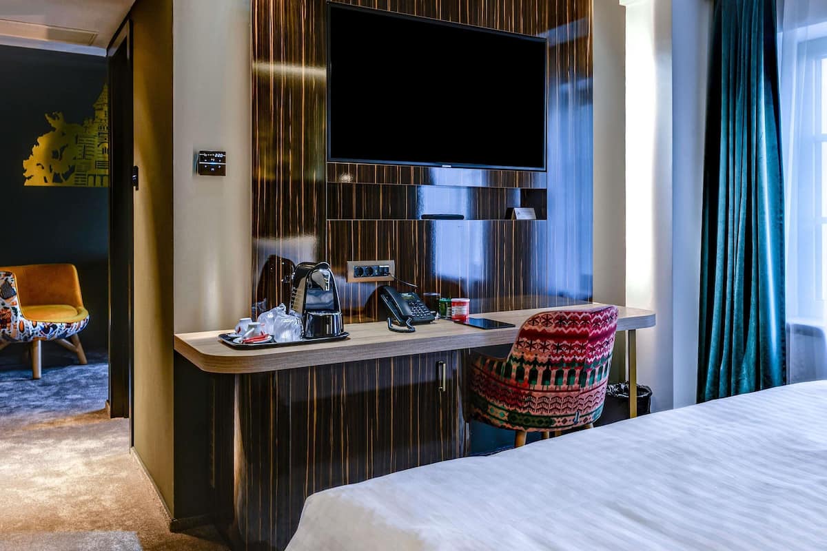 Habitación de hotel con cama, escritorio con cafetera, teléfono, televisor y una silla estampada.