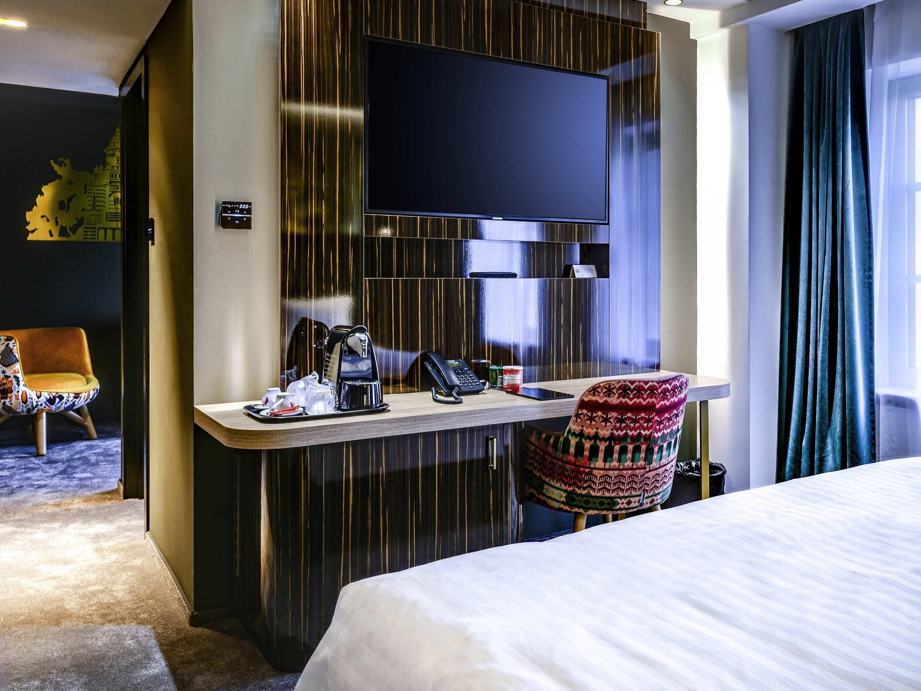 A hotel room with a bed, a desk with a telephone, a coffee maker, and a TV mounted on the wall.