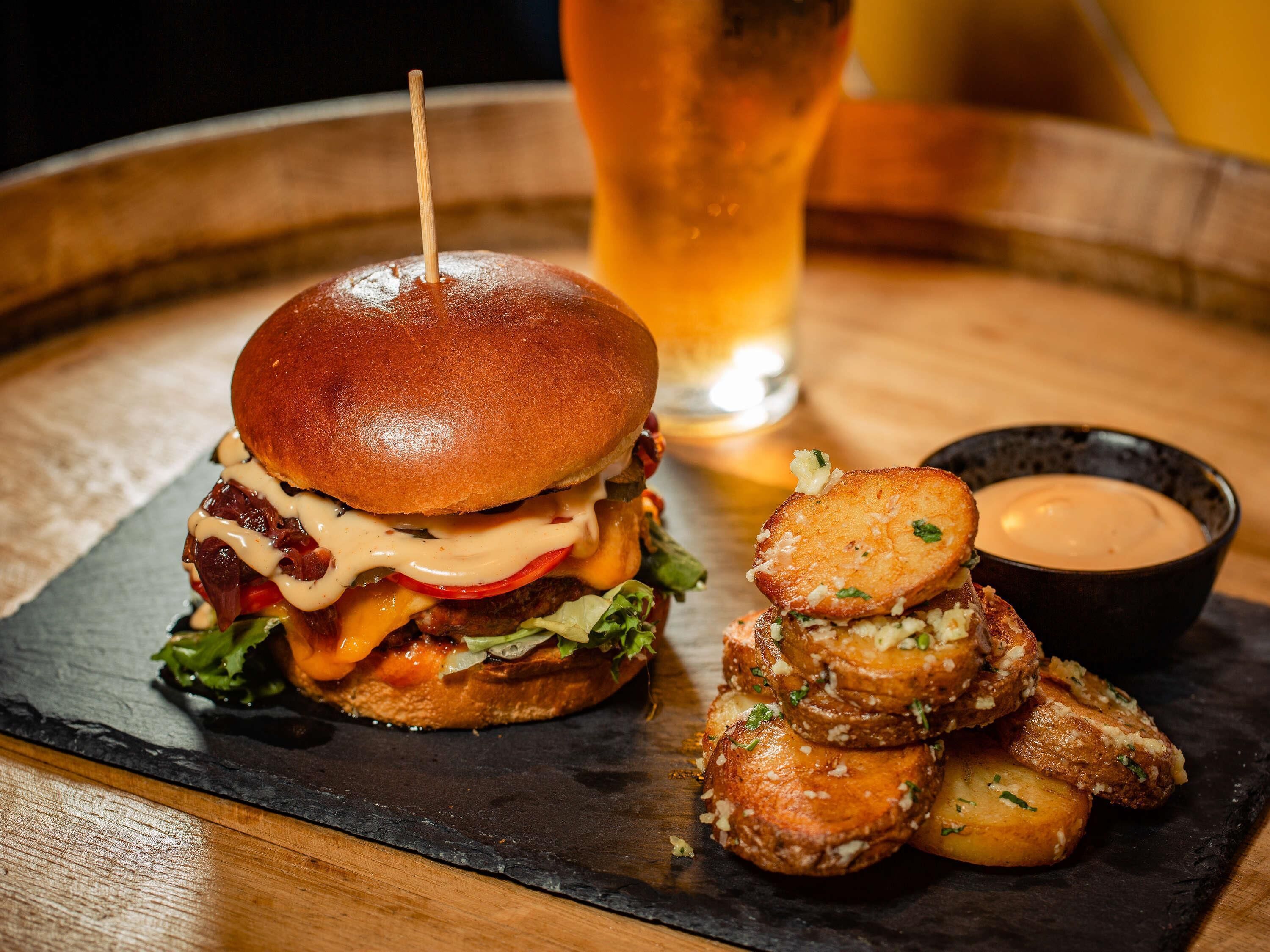 Una hamburguesa con un palillo, un vaso de cerveza y un cuenco de salsa sobre una tabla de pizarra.