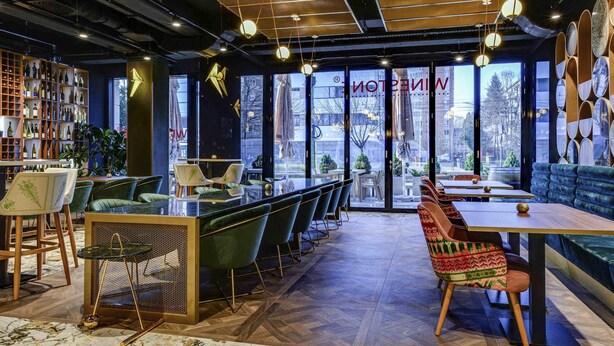 A modern restaurant interior with a bar area, seating, and a view through large windows. - Mercure Timisoara (Timisoara)