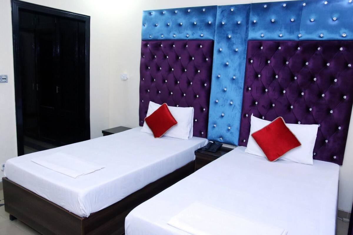 Habitación de hotel con dos camas individuales, cada una con almohada roja y ropa blanca. Los cabeceros están acolchados con una combinación de colores azul y morado.