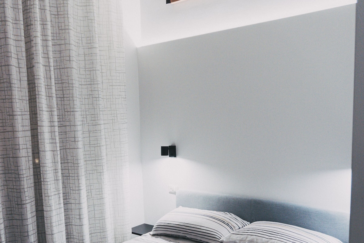 Una cama bien hecha con ropa de cama a rayas y toallas enrolladas, un cabecero y una lámpara fijada a la pared.