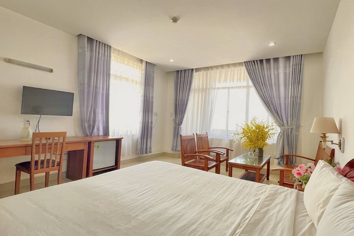 A hotel room with a bed, a desk with a TV, two chairs, a small table with flowers, and large windows with curtains.