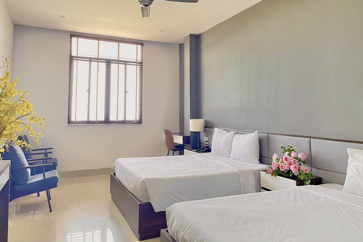 A hotel room with two beds, a desk, a chair, a ceiling fan, and a window with blinds.