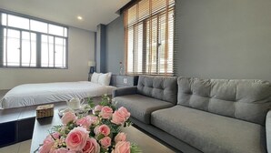 A modern hotel room with a grey sofa, a bed with white bedding, a small table with a vase of pink flowers, and large windows with blinds. - My Khe 2 Da Nang Seafront Hotel (Da Nang)