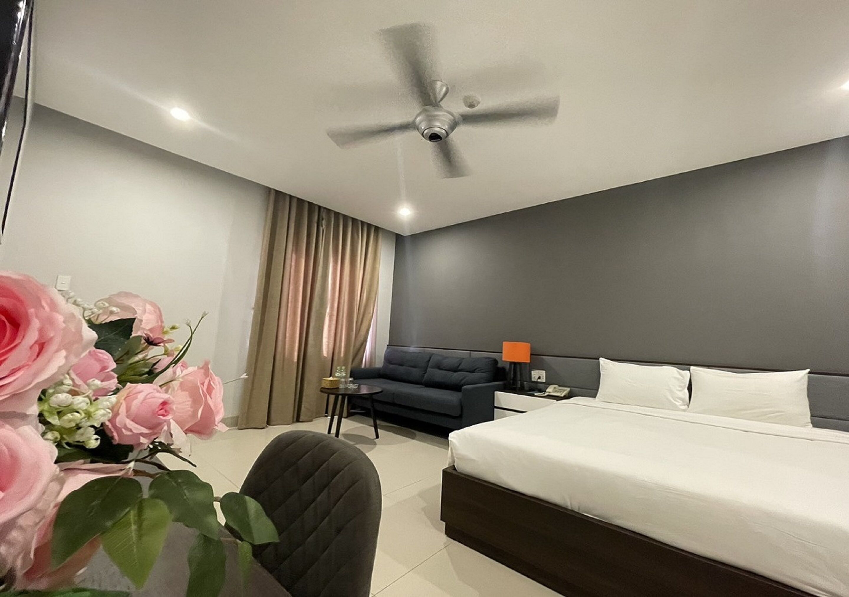 A modern hotel room with a bed, a sofa, a small table, and a ceiling fan.