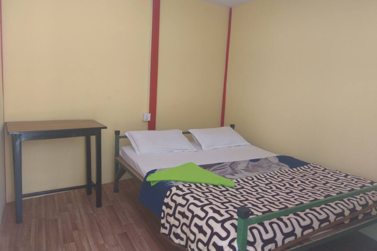 A small, single-bed room with a wooden floor, a bedside table, and a patterned bedspread.