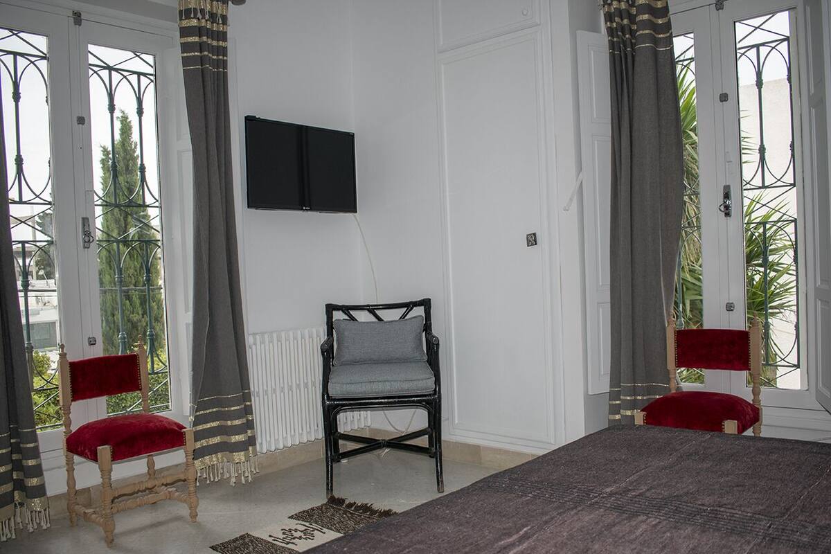 A room with two red upholstered chairs, a grey upholstered chair, a flat-screen TV on the wall, and a large window with curtains.