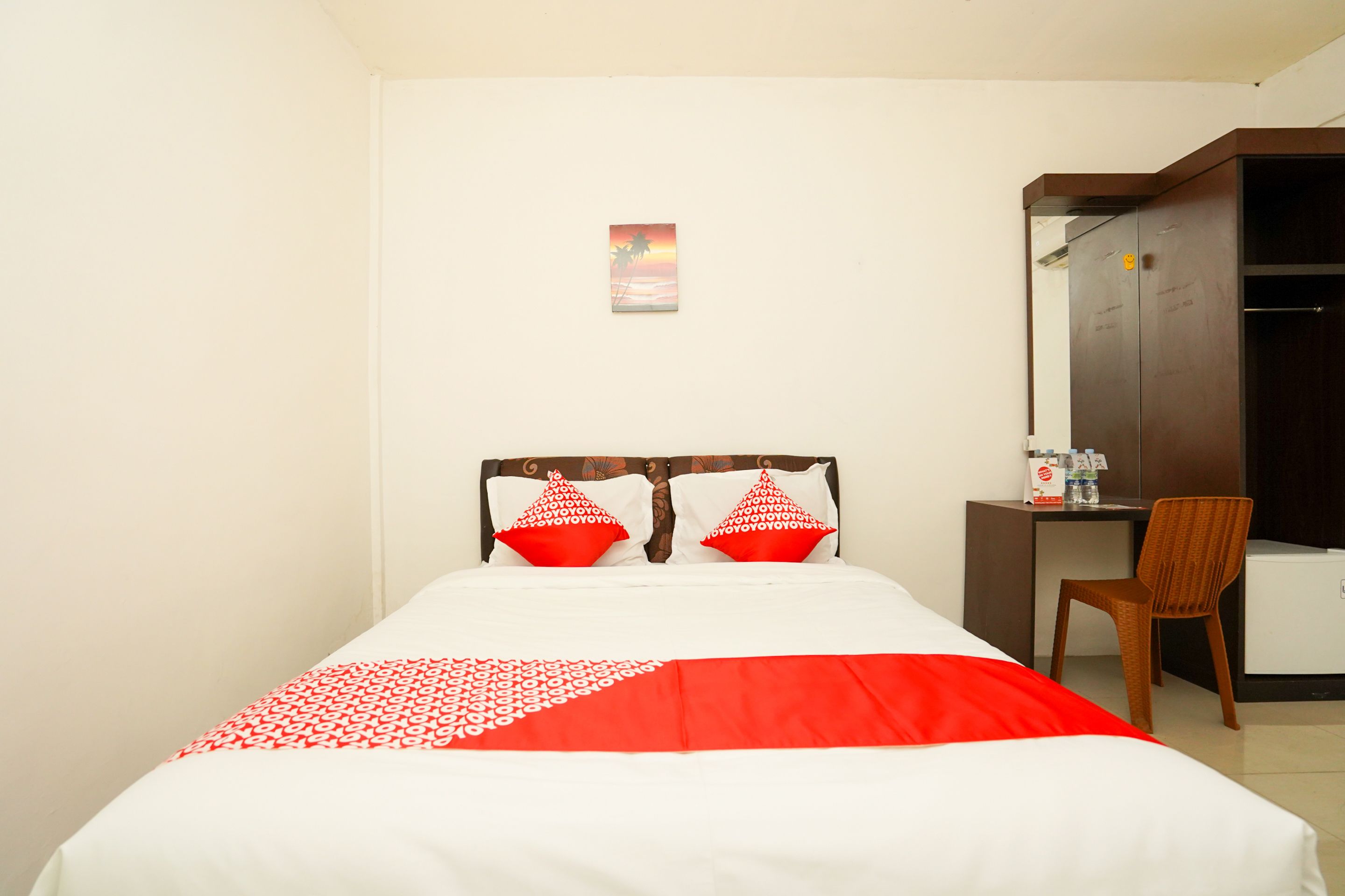 A hotel room with a bed, two red and white pillows, a small desk with a chair, and a wall-mounted picture.