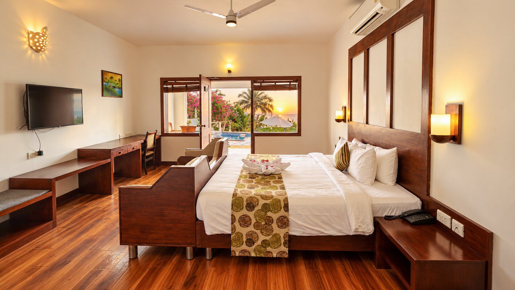 Habitación de hotel con cama, mesitas de noche, escritorio, silla, televisión y ventana con vista a una piscina y palmeras.