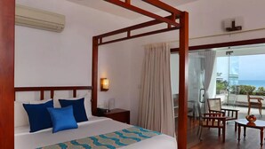 Una cama con dosel, almohadas azules, cabecera de madera y colcha estampada. Se observa una mesita de noche con lámpara. La habitación tiene un ventanal con cortinas y un balcón con muebles y una mesa de madera.