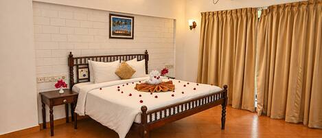 Una cama bien hecha con una colcha blanca, cabecera de madera y un cojín decorativo. Se observa una mesita de noche con un jarrón de flores y un equipo de aire acondicionado instalado en la pared.