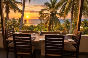 Mesa de comedor para dos con vista al atardecer sobre el océano, rodeada de palmeras.
