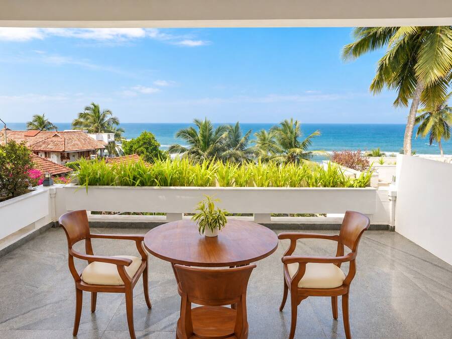 Een balkon met een houten tafel en stoelen, met uitzicht op de kust, palmbomen en de zee.