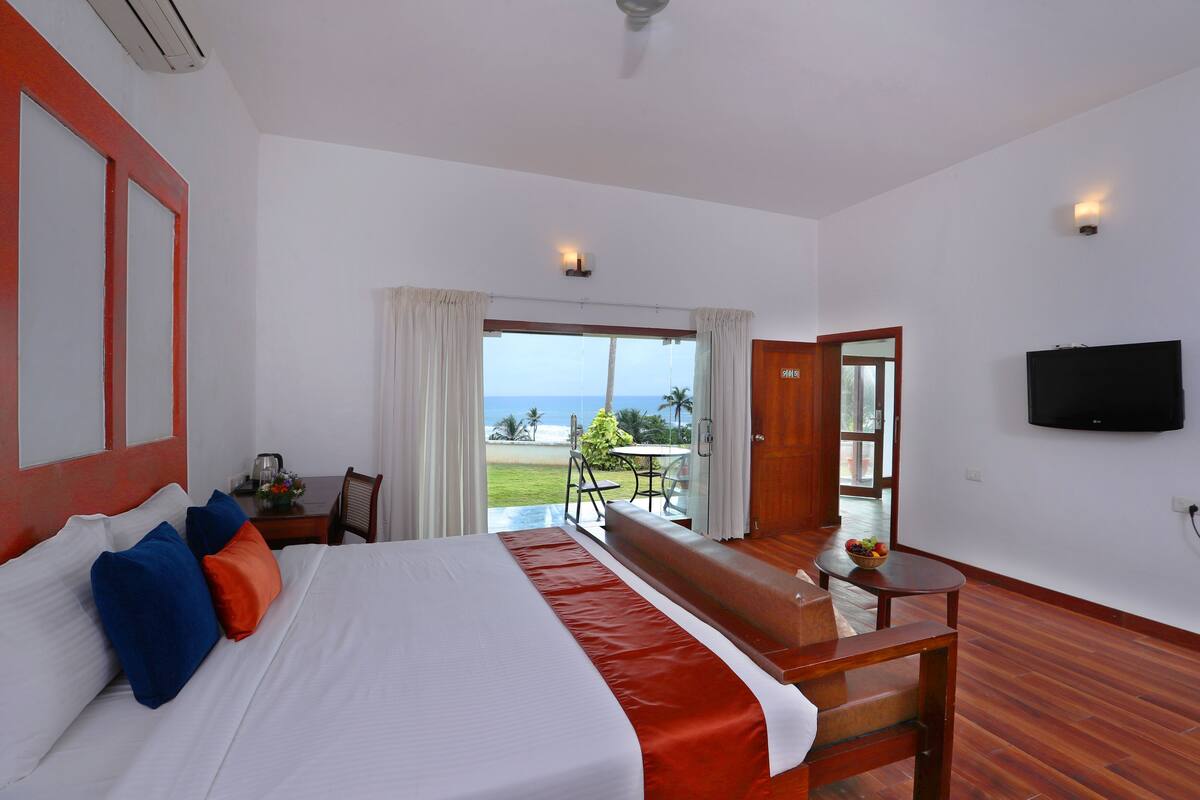 Ein Hotelzimmer mit einem großen Bett, einem Fernseher, einem Balkon mit Blick auf den Strand und einer Essecke.