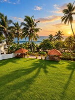Un resort tropical con dos estructuras de techo de paja, un césped verde exuberante, palmeras y vista al océano al atardecer.