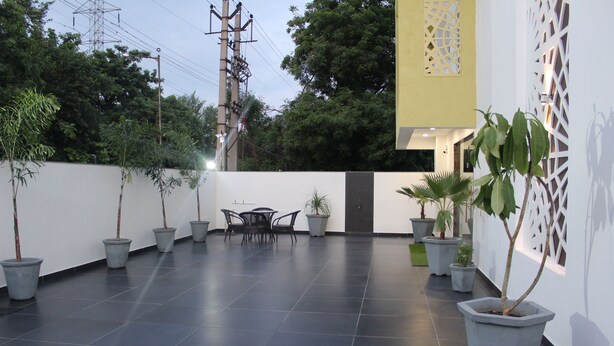 A spacious outdoor area with a tiled floor, potted plants, a table with chairs, and a white wall with decorative cutouts. - Hotel Sai (Bawal)