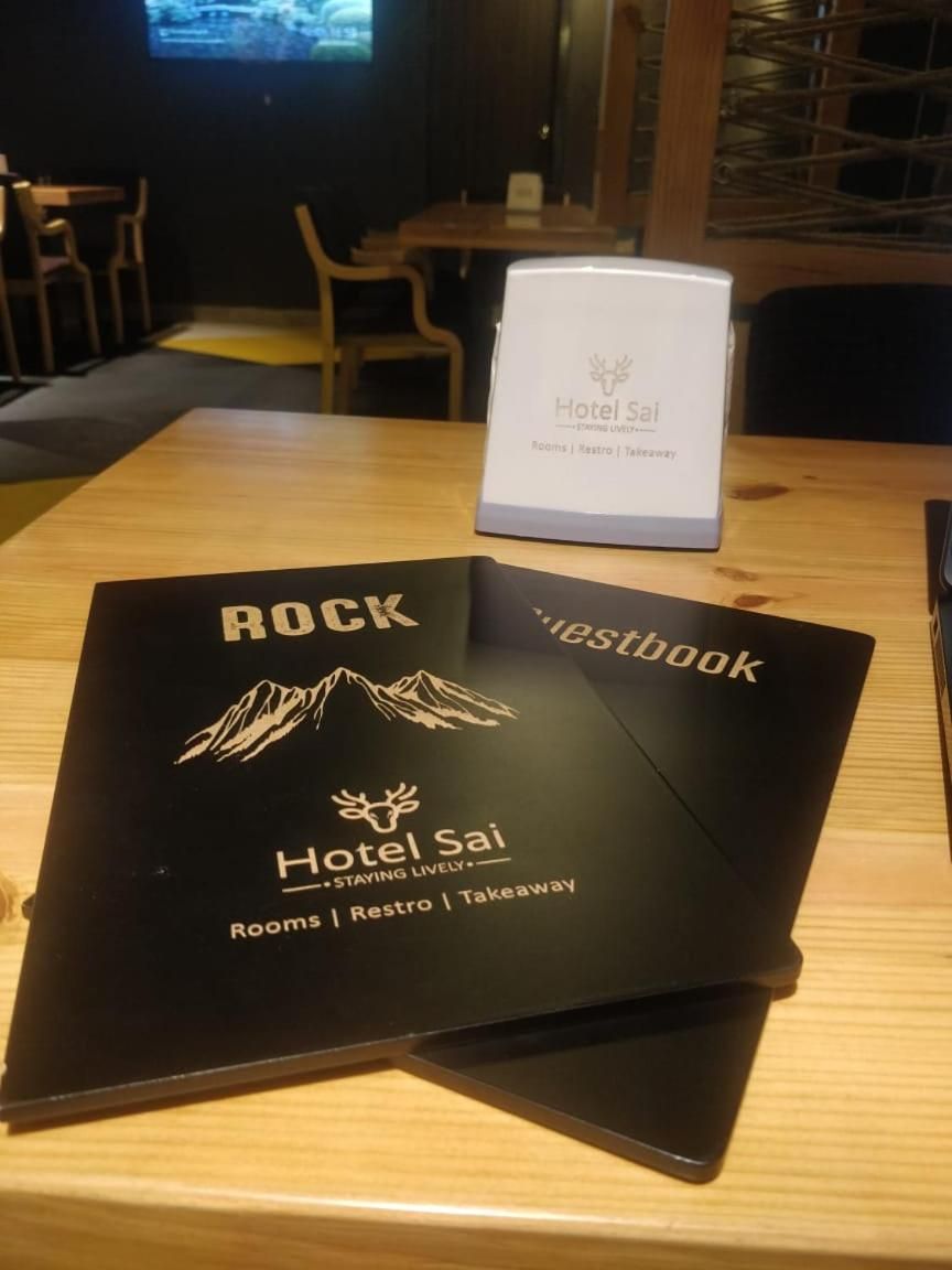 A black booklet titled 'Hotel Sai' with 'Rooms | Restro | Takeaway' on a wooden table.
