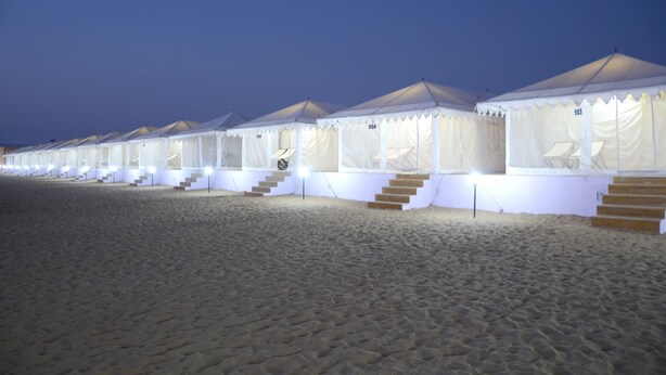 Une rangée de tentes blanches, chacune dotée de quelques marches menant à l’entrée, est disposée sur une plage de sable au crépuscule.