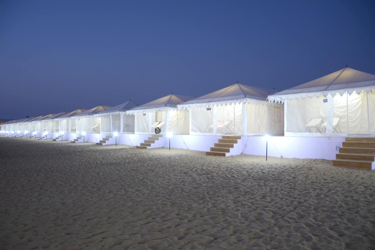 A row of white tents with steps leading up to the entrance, arranged on a sandy beach at dusk.