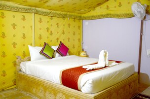A hotel room with a bed, pillows, a bedside table, a lamp, and a standing fan.
