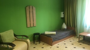 Une pièce avec un mur vert, un lit recouvert d’une couvre-lit à motifs, un fauteuil vert, un petit tabouret et une table avec une lampe.