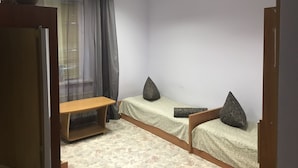 Une chambre avec deux lits simples, un banc en bois, une plante en pot et une fenêtre ornée de rideaux légers.