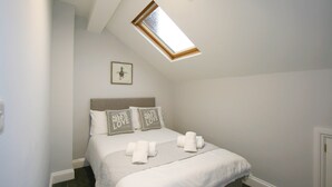 Quarto com teto inclinado, claraboia, cama com roupa de cama branca, dois travesseiros decorativos e um quadro na parede.