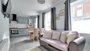 Uma sala de estar moderna com um sofá cinzento, uma cadeira de madeira e um tapete branco. Ao fundo, vê-se uma cozinha com armários brancos e uma janela com cortinas cinzentas.